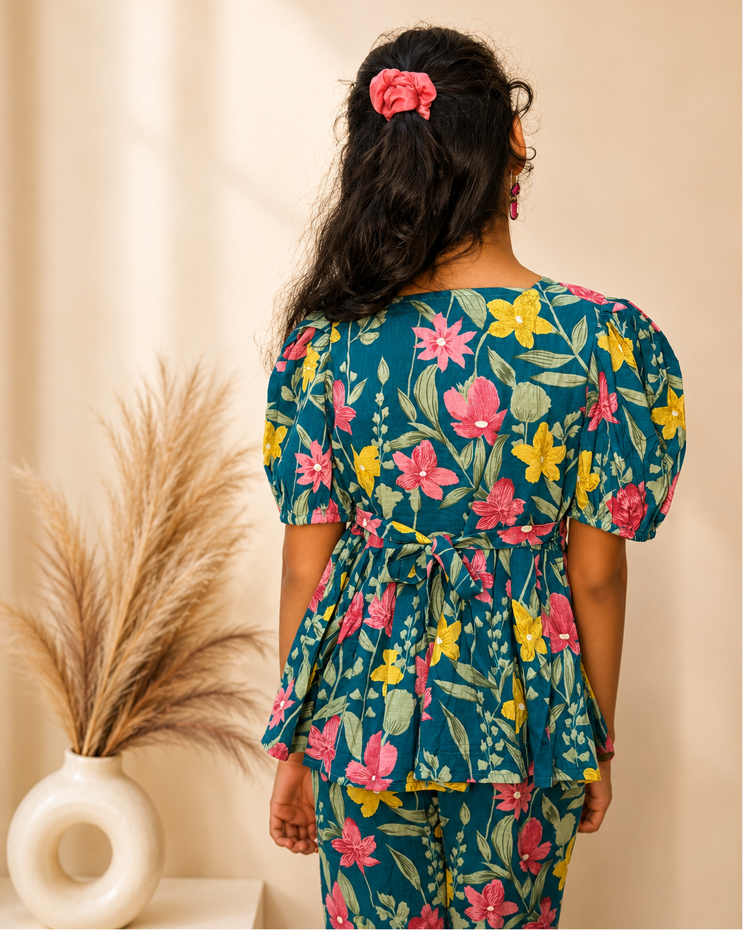 A woman viewed from behind wearing a teal floral peplum top and matching pants. The outfit features a vibrant pink, yellow, and green floral pattern.