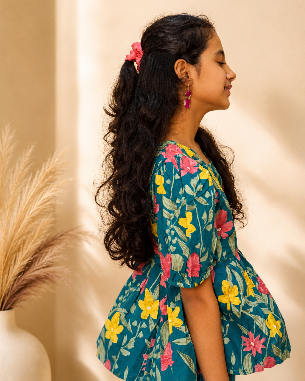 A young girl with long, wavy black hair tied back with a pink hair tie is shown in profile wearing a teal short-sleeve dress with a colorful floral print. She is standing in front of a warm beige wall with soft shadows, next to a white vase with dried pampas grass.