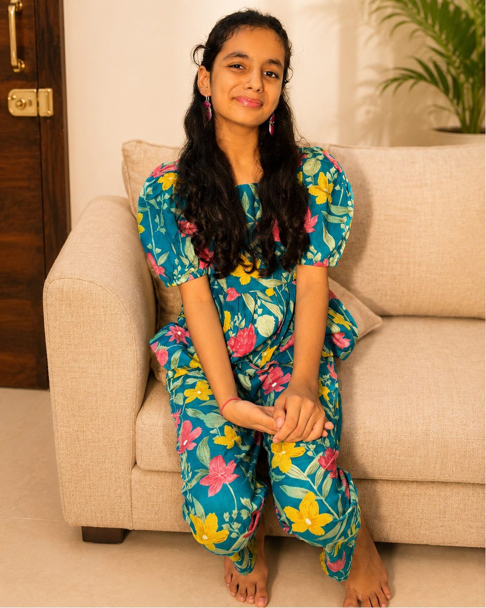 A young girl with long black hair sits barefoot on a beige fabric sofa, wearing a bright blue floral pajama set with pink, yellow, and green flowers. A dark wooden door is on the left, and a green potted plant is on the right.