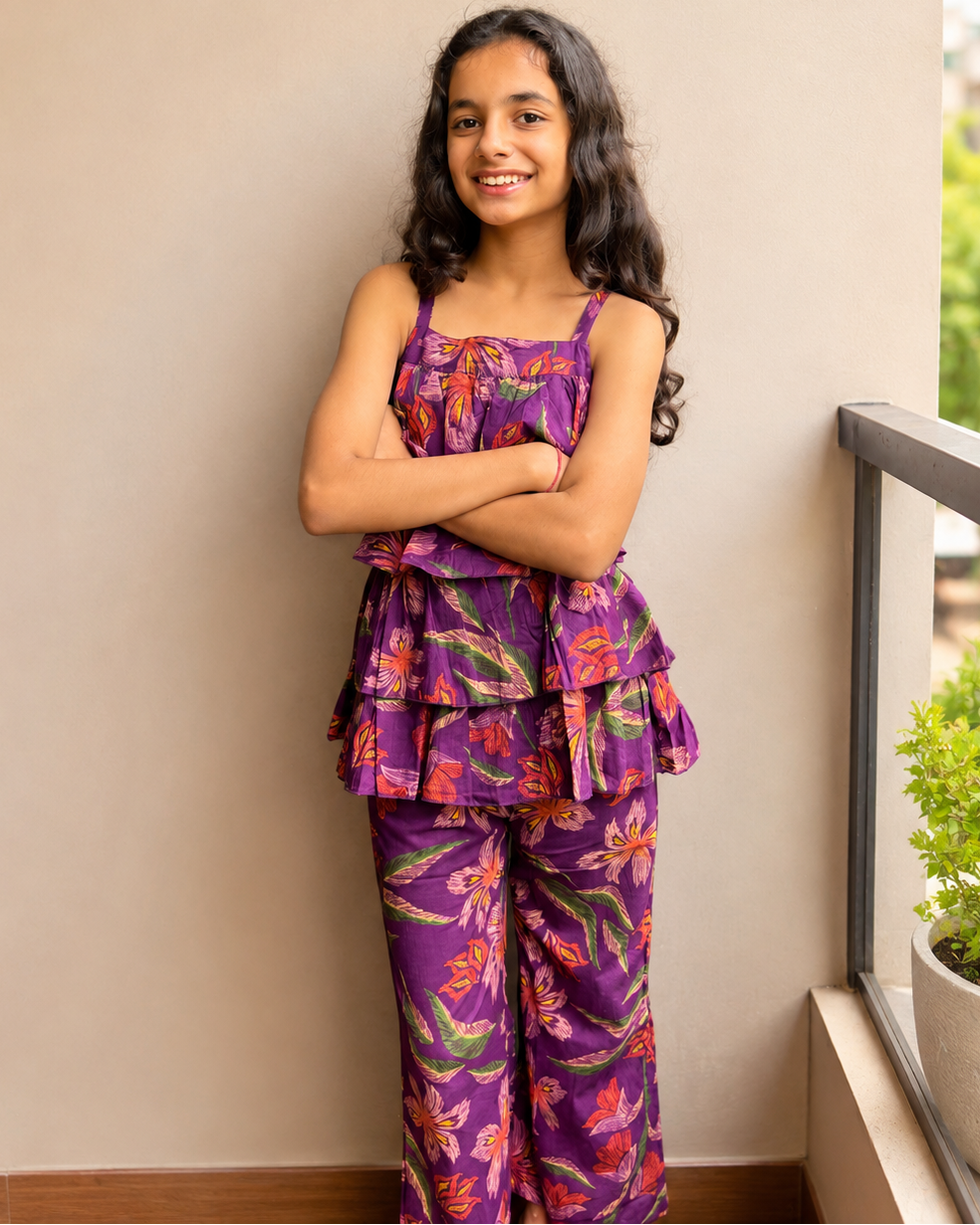 A young girl with long dark hair stands on a balcony wearing a purple two-piece pajama set with a colorful floral print. The set includes a sleeveless top with thin straps and a matching pair of wide-leg pants.