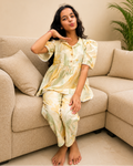 A young girl with long dark hair sits barefoot on a beige sectional sofa, wearing a matching short-sleeve button-front top and pants set in a soft yellow, cream, and green abstract print.
